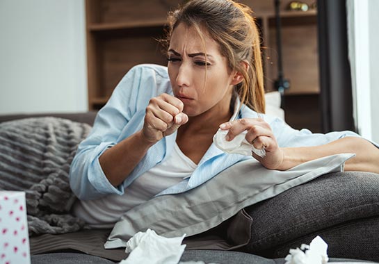 Mulher deitada no sofá, tapada com cobertor, tossindo, visivelmente com uma doença semelhante a uma gripe - Clínica do Pulmão