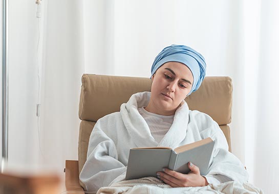 Mulher branca com lenço enrolado na cabeça e vestida com roupão branco, sentada em uma cadeira de tratamento para quimioterapia, lendo um livro - Clínica do Pulmão